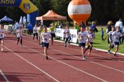 Bieżnia na stadionie. Biegną tu chłopcy w sportowych strojach. Nad bieżnią jest dmuchana brama z napisem Ciechanów i herbem miasta. Po prawej fragment murawy stadionu. Jest tu sporo dorosłych i dzieci, zadaszone stoisko i balon z logo miasta. W tle drzewa