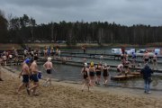 Pogoda jest szara i smutna. Na drzewach liściastych w lesie po lewej nie ma liści. Na kąpielisku trwa zlot osób morsów. Morsujący ubrani są w kostiumy kąpielowe, na głowach mają czapki. Na nogach buty do morsowania. Wiele osób jest na pomostach i w wodzie