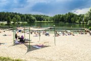 Zdjęcie zrobione od strony ulicy Sońskiej. Na pierwszym planie plaża z plażowiczami i żaglami przeciwsłonecznymi. Dalej w głąb woda. Na skraju przeciwległego brzegu akwenu bujna zieleń: szuwary, drzewa i krzewy. Na niebie kłębią się chmury.