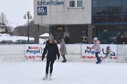 W kadrze fotografii znajdują się trzy osoby jeżdżące na łyżwach po zaśnieżonym lodowisku. Pada śnieg. Na hali sportowej dokładnie widać adres 17 Stycznia 60C. Na znajdującym się niżej termometrze 0 stopni Celsjusza.