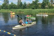 Na wodzie Pływają ludzie w trzech kajakach i na żółtym rowerze wodnym. Ludzie mają na sobie kapoki. Na brzegu akwenu rośnie wysoki, zielony tatarak, a dalej w głębi są drzewa.