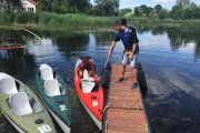 Na drewnianym pomoście przy brzegu stoi tu mężczyzna, który wiosłem przytrzymuje kajak, by pomóc drugiemu mężczyźnie wyjść na pomost. Po lewej stronie są przycumowane dwa puste kajaki. W oddali, na brzegu rośnie tatarak. Dalej po lewej jest biały budynek.