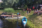 Na brzegu są dwa drewniane pomosty, każdy z pięcioma oponami zabezpieczającymi sprzęt przed uszkodzeniem podczas podpływania. Przy brzegu zacumowane są trzy kajaki, Na wzniesieniu na brzegu siedzi kilka osób i stoją też dwie osoby z wiosłami w rękach