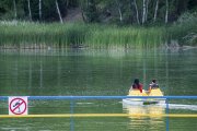 Na zdjęciu zbiornik wodny. Na dole kadru poręcz pomostu, na której zamocowany jest znak zakazujący skoków do wody. Za pomostem na rowerze wodnym płyną dwie osoby. Na przeciwległym brzegu akwenu są piękne zielone szuwary, a dalej wysoki las.