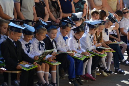 Mali uczniowie w odświętnych strojach siedzą w pierwszym rzędzie. Na kolanach kolorowe książki i kredki. W drugim rzędzie stoją dorośli.