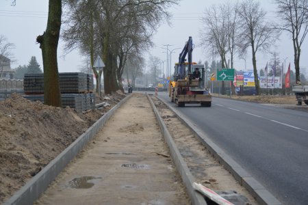 Pas ziemi ograniczony krawężnikami po obu stronach, przygotowany pod budowę chodnika. Po prawej jezdnia, po której jedzie duża maszyna do robót drogowych