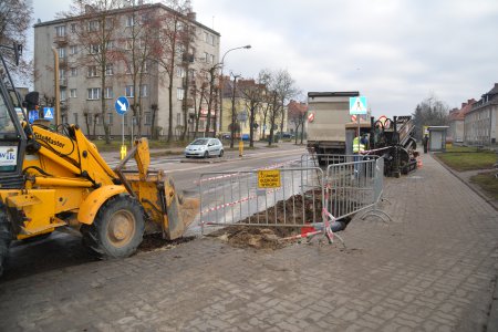 Rozkopany fragment chodnika, widać fragment podziemnej instalacji. Obok stoi koparka. W tle budynki wielorodzinne