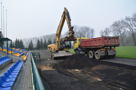 Trwa rozbiórka starej nawierzchni bieżni na miejskim stadionie. Pracuje koparka, która zrywa starą nawierzchnię. Obok stoi samochód z przyczepą, na który ładowany jest zdjęty żużel