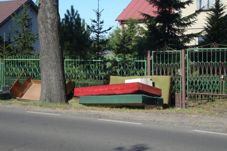 Przed bramą wjazdową na posesję stoją wystawione stare meble: kanapa i tapczanik. O ogrodzenie oparty jest materac