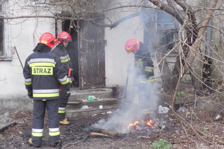 Trzech strażaków na miejscu zgłoszenia. Jeden z nich dogasza ognisko. W tle zrujnowany budynek, wokół dużo śmieci