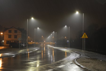 Zdjęcie po zmierzchu. Lampy uliczne oświetlają mokrą od deszczu ulicę. Lekka mgła powoduje, że światło jest łagodne, miękko skupione. Świetlne smugi odbijają się w mokrym asfalcie.