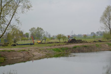 Nad brzegiem stawu, na niezagospodarowanym terenie widać maszyny wykonujące wykopy pod montaż urządzeń