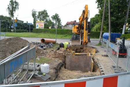 Plac budowy otoczony barierkami. Pracuje koparka. wykonuje wykop, w który montowane są zabezpieczenia zapobiegająe osypywaniu się ziemi ze ścian wykopu