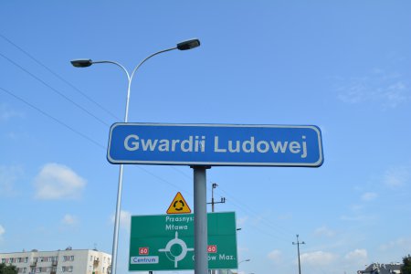 Oznakowanie z nazwą ulicy Gwardii Ludowej. W tle inne oznakowanie drogowe informuje o niedalekim rondzie. Za nimi fragment budynku wiekorodzinnego