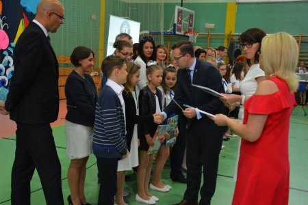 Prezydent Ciechanowa w towarzystwie Skarbnik urządu i nauczycieli gratuluje dzieciom osiągnięć w nauce i wręcza dyplomy. Dzieci stoją rzędem.