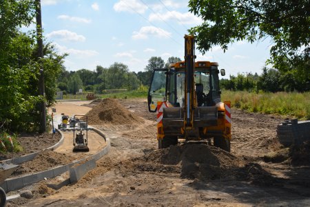 Plac budowy. Dużo żwiru. Pośrodku pracuje koparka., po lewej między ułożonymi krawężnikami ubijane jest podłoże pod nawierzchnię dojazdu