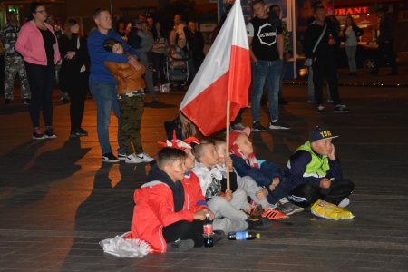 Kilku chłopców siedzi w rzędzie na posadzce pl. Jana Pawła II. Jeden z nich trzyma polską flagę narodową. W tle widać stojących ludzi, zwróconych w stronę ekranu