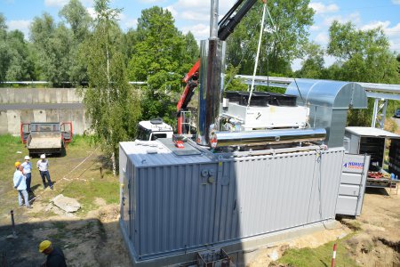 Plac budowy. Pośrodku prostokątny obiekt podobny do blaszanego garażu. Na tym kontenerze umiejscowiono dwie rury. Jedna ułożona poziomo, druga ustawiona pionowo. Za kontenerem samochód z podnośnikiem, który na dachu obiektu ustawia czworoboczny element z czterema okrągłymi otworami