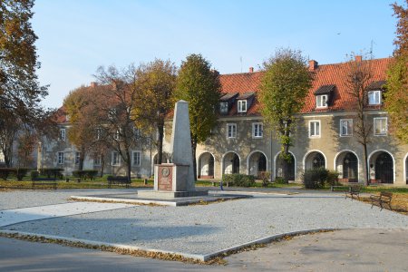 Plac Piłsudskiego. Na pierwszym planie kolumna upamiętniająca Marszałka, w tle krzywa hala. Między pomnikiem i krzywą halą w jesiennych barwach