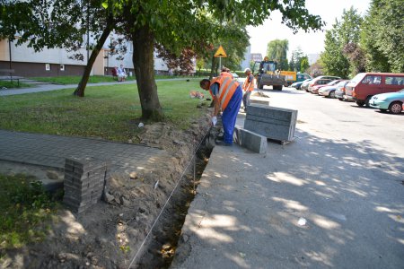 Wzdłuż jezdni robotnik wykopuje wąski dołek. Za jego plecami stoi słupek ustawionych jedna na drugiej płyt krawężnikowych