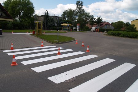 Świeżo namalowane pasy na przejściu dla pieszych. W części, która jeszcze nie wyschła stoją niewysokie słupki w kształcie stożka uniemożliwiające wjazd na tę część pasów