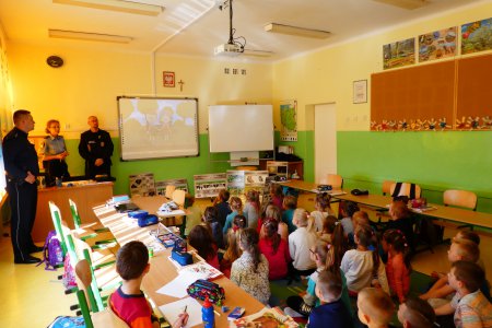 Zdjęcie zrobione podczas zajęć w szkolnej sali. Pośrodku sali między dwoma rzędami ławek, na podłodze siedzą uczniowie. Przy tablicy stoją: strażniczka miejska, strażnik i policjant