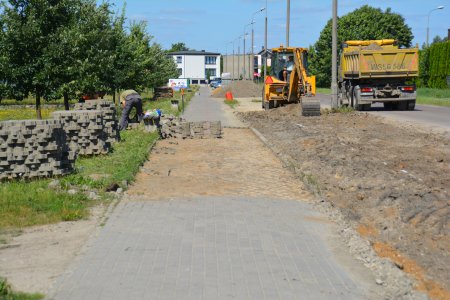 Pośrodku zdjęcia długi chodnik, którego fragment  jest właśnie układany. Po lewej ułożone na trawniku duże kupki brukowej kostki. Po prawej, na jezdni stoi koparka i wywrotka. Szeroki pas między chodnikiem a jezdnią jest pozbawiony trawy i wyrównany pod budowę parkingu