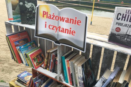 Regał, na którym stoją książki. Na regale umieszczona jest tabliczka w kształcie książki a na niej napis 