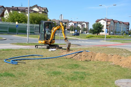 Na dużym trawniku przy ul. Armii Krajowej niewielka koparka z wąską łyżką kopie długi dołek, w którym będzie położony kabel. Gruby kabel elektroenergetyczny leży przygotowany obok  na trawie