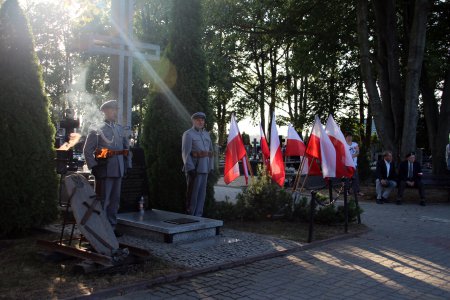 Harcerska warta przy nagrobku z wysokim krzyżem. Obok zamontowano wiele flag w barwach narodowych. W tle wysokie drzewa. Przez konary drzew przedziera się jasny promień słońca