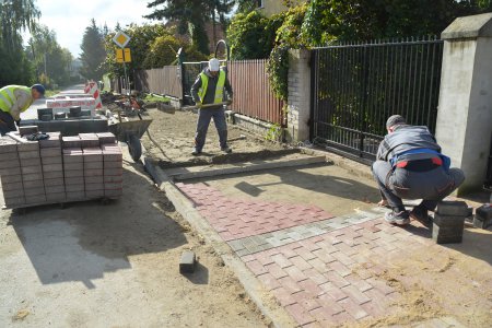 Trwa układanie chodnika z betonowej kostki. Jeden z pracowników wyrównuje grunt, a drugi układa kostkę. Obok powstającego chodnika ułożone palety z kostką