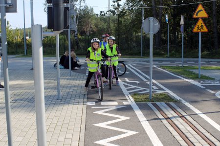 Dzieci na rowerach korzystają ze ścieżki rowerowejścieżce rowerowej. Mają na sobie kamizelko odblaskowe, na głowach kaski ochronne