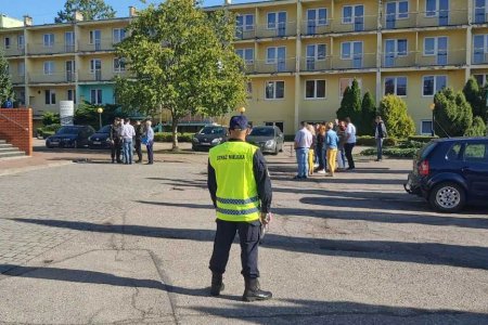 Na pierwszym planie zdjęcia stoi strażnik miejski odwrócony plecami do fotografującego. Ma na sobie żółtą kamizelkę z napisem Straż Miejska. W głębi stoją dwie grupki ludzi, a w tle znajduje się Hotel Olimpijski.