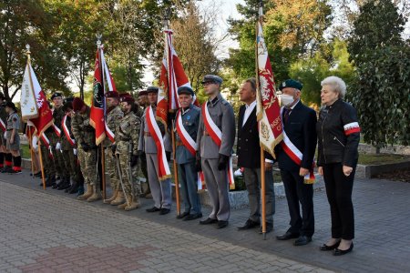 W rzędzie stoją mundurowi różnych formacji z biało czerwonymi szarfami włożonymi na skos oraz opaskami na rękawach. Kilku z żołnierzy trzyma nasze narodowe flagi. W tle drzewa i krzewy.