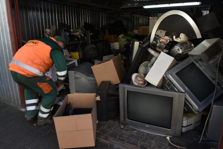 Duże pomieszczenie ze ścianami z blachy. Wypełnione jest  różnymi urządzeniami, między innymi telewizorami, monitorami komputerów, odbiornikami radiowymi i odtwarzaczami płyt CD. W wejściu pracownik w pomarańczowej kamizelce schyla się do jednego ze sprzętów.