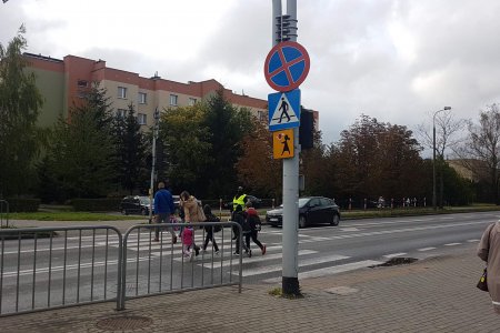 Przejście dla pieszych. Po lewej barierki ochronne i słup, na którym są znaki drogowe. Na przejściu znajduje się kilkoro dzieci i dwoje dorosłych. Ruchem kieruje Strażnik Miejski, stojący obok pasów. W oddali szkoła.