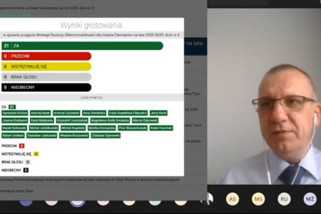 Kadr z zapisu wideo sesji Rady Miasta. Po lewej na białym tle wyniki głosowania pokazane jako poziome paski w różnych kolorach. Od góry napis: Wyniki głosowania w sprawie przyjęcia Strategii Elektromobilności. Niżej graficznie pokazane wyniki głosowania. Od góry leżą: najdłuższy zielony pasek z napisem 21 za, czerwony - zero przeciw, żółty - zero wstrzymujących się, szary - zero brak głosu, czarny - zero nieobecny. Niżej na zielono zaznaczone nazwiska wszystkich radnych. Po prawej widać głowę i fragment klatki piersiowej przewodniczącego Rady Miasta. Ma okulary, białą koszulę i krawat. Włosy króciutko ostrzyżone.