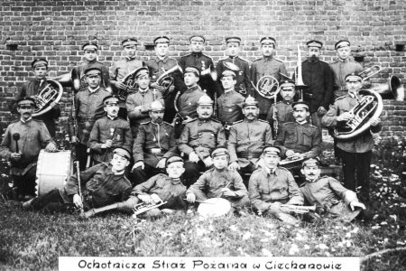 Stare, czarno-białe zdjęcie orkiestry Ochotniczej Straży Pożarnej z 1915 roku. Informuje o tym napis na dole fotografii. Na zdjęciu, na tle ceglanego muru, duża grupa mężczyzn w mundurach z instrumentami. Są tu różne instrumenty dęte: m. in. trąbka, puzon, waltornia, a także duży bęben oraz werbel.