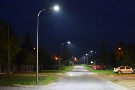 Zdjęcie zrobione po zmierzchu. Po obu stronach utwardzonej drogi stoją rzędy lamp ulicznych. Lampy się palą. Po obu stronach drogi są zadrzewione prywatne posesje. Po prawej przy posesjach parkują dwa auta.