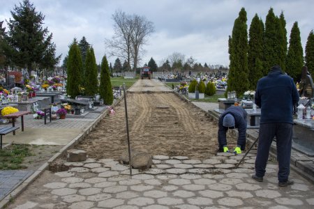 Na zdjęciu szeroka cmentarna aleja. Po jej obu stronach są nagrobki. Na grobach wiele kolorowych chryzantem. Dwóch pracowników usuwa grube betonowe płyty przy pomocy długich, solidnych prętów. W oddali, na nowej nawierzchni stoi ciągnik. Między starą i nową częścią jest kilkumetrowy pas gołej ziemi.