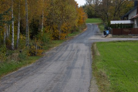 Malowniczo położona droga. Po lewej jesienne brzozy oraz inne drzewa i krzewy w kolorach żółtym, zielonym i rudym. Po prawej fragment łąki, za nią droga i ogrodzona posesja. U szczytu droga skręca w lewo. W głębi widać łąkę, a za nią drzewa.