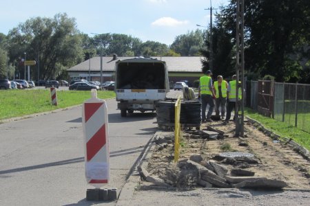 Wąska część ulicy Tatarskiej prowadzącej do parkingu przed sklepem Biedronka. Na jezdni stoi samochód dostawczy. Po prawej stronie jezdni rozbita część nawierzchni chodnika. Leżą tu betonowy gruz. Dalej stoi duża paka brukowej kostki owinięta folią. Obok znajduje się czterech robotników w odblaskowych kamizelkach.