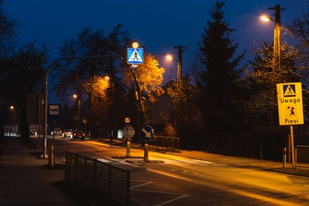 Zdjęcie wykonane po zmroku. Na zdjęciu dobrze oświetlone przejście dla pieszych. Przed przejściem po prawej stronie ulicy oraz nad przejściem są znaki drogowe informujące o przejściu dla pieszych.