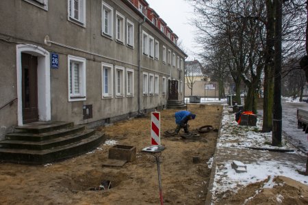 Wzdłuż bloku widać szeroki pas gruntu, z którego zdjęto już stary chodnik. Pracuje tu mężczyzna, który jest pochylony nad studzienką kanalizacyjną. W pobliżu studzienki znajduje się biało czerwony słupek ostrzegawczy.