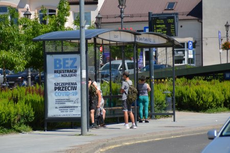 Przystanek autobusowy z przeszkloną wiatą. Stoi tu kilka osób. Na bocznej ścianie wiaty umieszczono plakat zachęcający do szczepień.