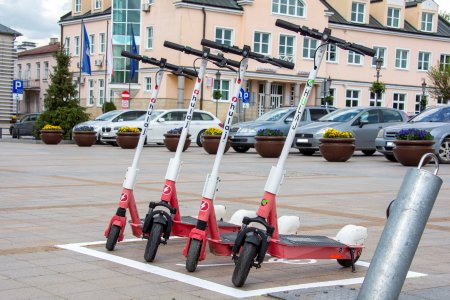 Cztery elektryczne hulajnogi stoją na placu Jana Pawła drugiego, w prostokątnym polu otoczonym grubą białą linią. W tle stoi bank, a w głębi po lewej znajduje się fragmencik budynku Urzędu Stanu Cywilnego przy ratuszu.