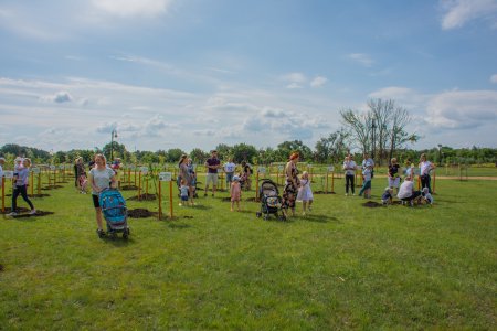 Na dużym terenie porośniętym trawą rosną młode drzewka. Obok drzewek stoją młodzi ludzie z małymi dziećmi w wieku około 2-3 lat, gdzieniegdzie obok rodziców stoją wózki dziecięce spacerówki.