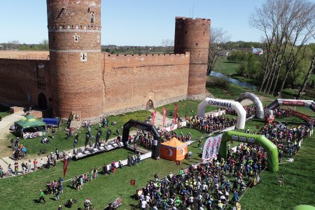 Zamkowe błonia i Zamek Książąt Mazowieckich widziany z wysoka. Na błoniach trwają zawody. Podłużne strefy wzdłuż południowej ściany zamku wyznaczono linami. Strefa bliżej warowni jest zabezpieczona białymi bandami. Nad obiema strefami stoją dmuchane bramy, Strefa bliżej rzeki jest pełna ludzi.