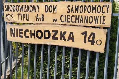 Na ogrodzeniu z metalowych prętów wiszą dwie podłużne zawieszone w poprzek tablice. Na tej wyżej znajduje się napis dużymi literami: Środowiskowy Dom Samopomocy typu A i B w Ciechanowie. Na niższej tablicy jest napis: Niechodzka 14o.