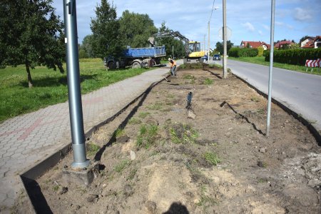 Szeroki pas ziemi między chodnikiem i jezdnią. W oddali koparka zrywa wierzchnią warstwę gruntu wraz z chwastami i wrzuca ją do wywrotki.